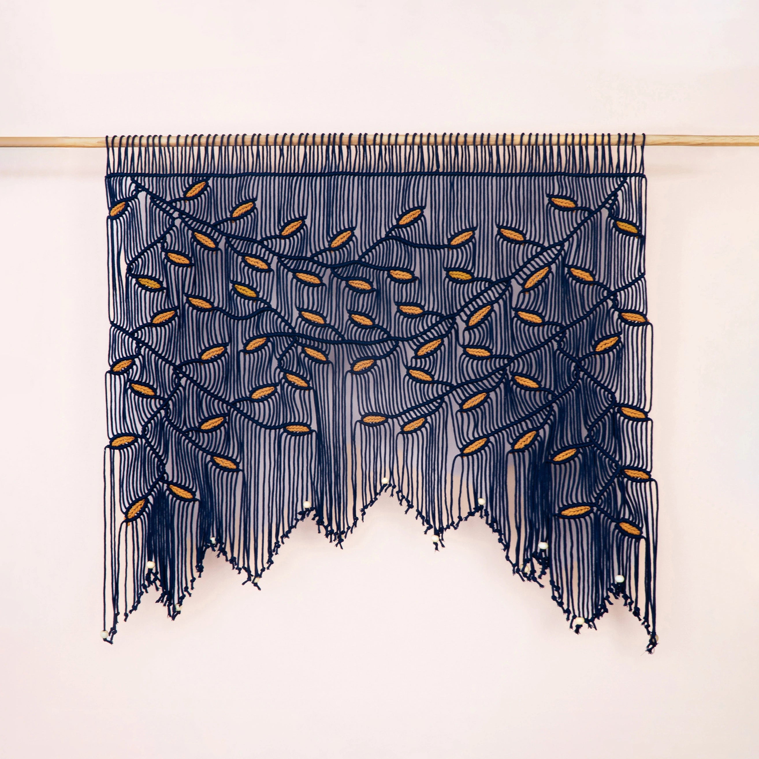 A bohemian-style wall hanging macrame wall tapestry with a tree branch pattern, featuring dark navy blue cords and small gold leaf accents, suspended from a light wood rod against a plain off-white wall.